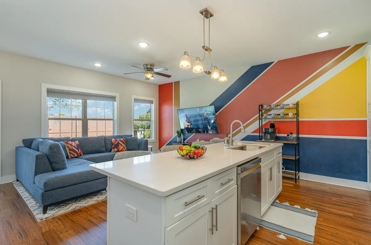 This image captures an open-concept living area featuring a comfortable sofa facing a kitchen island. A colorful accent wall and a television are visible, with plenty of natural light filling the space. The setup highlights both the living and kitchen areas working together, reinforcing the sense of connectedness and artistic flair.