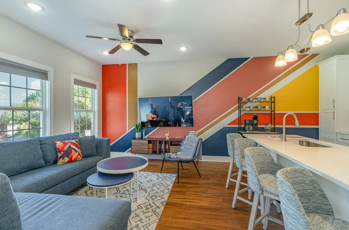 The image showcases a cheerful, designer-inspired living space with a bold colorful accent wall, inviting modern sofas arranged for conversation or relaxation, and a large TV setup perfect for movie nights. The area feels spacious yet cozy, hinting at opportunities for gatherings with friends or family.