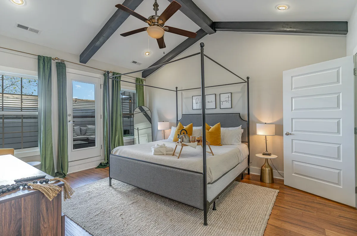A calm and welcoming bedroom featuring a four-poster bed with yellow accent pillows, large mirror, green curtains, and a wooden dresser. The scene evokes a sense of peace and comfort, ideal for a primary bedroom retreat.