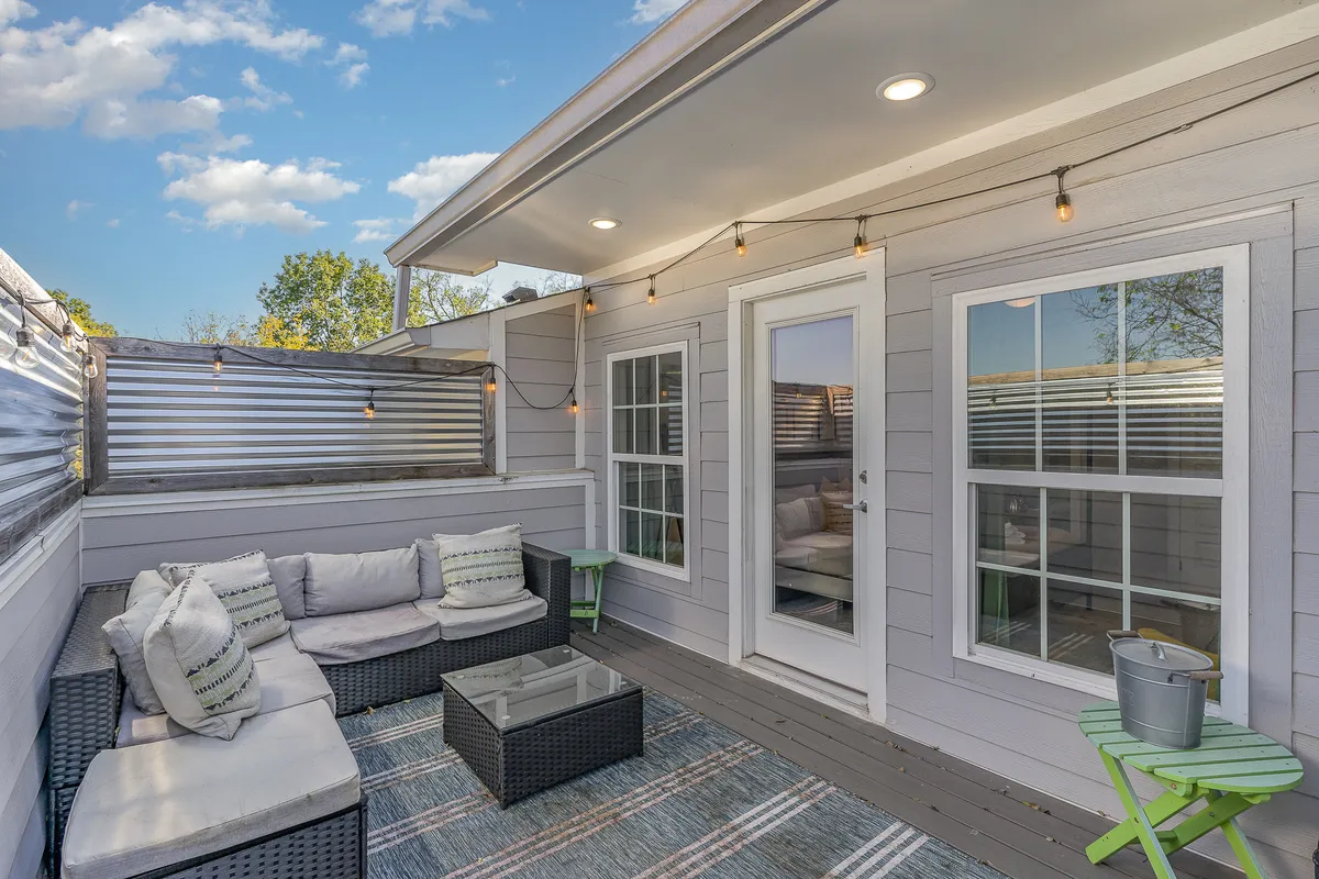 An inviting outdoor seating area illuminated by string lights, featuring a glass coffee table and comfortable chairs. The setting is styled for relaxation or gatherings, with lush greenery visible in the background and a cozy, welcoming mood.