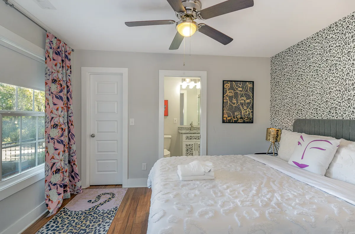 A stylish bedroom adorned with decorative pillows, colorful curtains, stylish artwork above the bed, and a vanity mirror. The space is bathed in natural light and exudes both comfort and artistic flair.