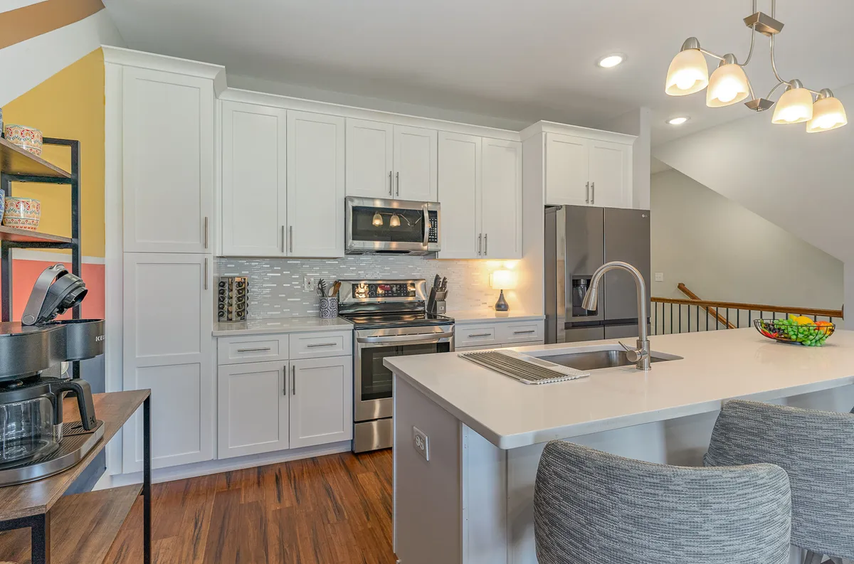 A bright and cheerful kitchen scene with a central island, stainless steel appliances, open shelving adorned with decorative items, and a colorful accent wall. The space is clearly designed for entertaining and group gatherings, with bar seating that encourages conversation and togetherness.