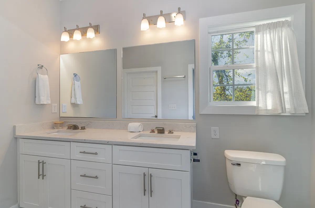 A double sink vanity with large mirrors is flooded with natural light from a nearby window. The bathroom gives off a serene, spa-like feel, highlighting how the home’s bath essentials and details are curated for guest comfort and relaxation.